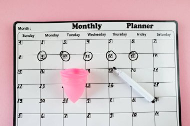 Beyaz planlayıcı ve açık pembe arka planda adet bardağı. Pastel renk. Yakın plan. Adet döngüsü.