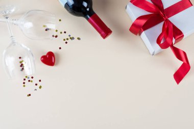 Sevgililer Günü konsepti, beyaz fotoğraf çerçevesi, iki kadeh şarap, hediye kutusu, bir şişe şarap, bej arka planda kalp. Boşluğu kopyala, metin için yer.