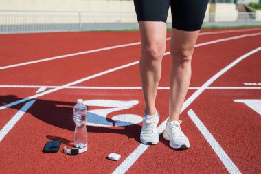 Spor ayakkabılı kadın ayakları, yeniden kullanılabilir cam su şişesi, kulaklık ve kırmızı spor halısında akıllı telefon. Güneşli bir günde aktif eğitim için spor su dengesi kavramı. Sıcak yaz.