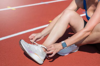 Spor ayakkabı bağcıklarını eğitirken bağlayan kadın. Spor kıyafetli bir kadın ayakkabı bağı bağlayıp çömeliyor. Açık hava eğitim stadyumunda gri gardırop ve beyaz spor ayakkabılar, kadın vücudunun yakın çekimi.