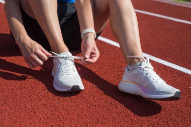 Spor giyinen, çömelmiş ve ayakkabı bağı bağlayan bir kadının yakın çekimi. Antrenman sırasında kadın ayakkabı bağcıkları spor. Moda kıyafetleri ve açık hava antrenman stadyumunda parlak ayakkabılar.