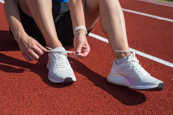 Spor giyinen, çömelmiş ve ayakkabı bağı bağlayan bir kadının yakın çekimi. Antrenman sırasında kadın ayakkabı bağcıkları spor. Moda kıyafetleri ve açık hava antrenman stadyumunda parlak ayakkabılar.