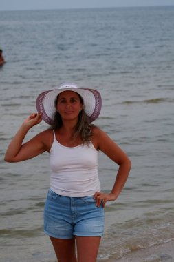 Tatil kumsalındaki kadın fotoğraf çekimi yapıyor farklı pozlar ve mekanlarda gülümsüyor sarı saçlı hasır şapka farklı çekim seçenekleri sunuyor güneşlenmeler deniz ve kumda yürüyor.