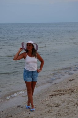 Tatil kumsalındaki kadın fotoğraf çekimi yapıyor farklı pozlar ve mekanlarda gülümsüyor sarı saçlı hasır şapka farklı çekim seçenekleri sunuyor güneşlenmeler deniz ve kumda yürüyor.