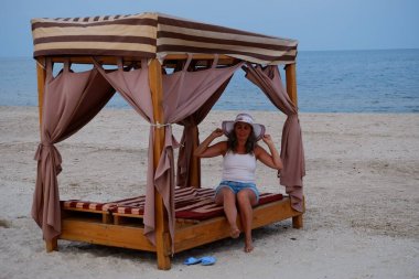 Tatil kumsalındaki kadın fotoğraf çekimi yapıyor farklı pozlar ve mekanlarda gülümsüyor sarı saçlı hasır şapka farklı çekim seçenekleri sunuyor güneşlenmeler deniz ve kumda yürüyor.