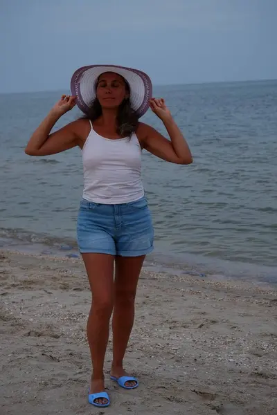 Tatil kumsalındaki kadın fotoğraf çekimi yapıyor farklı pozlar ve mekanlarda gülümsüyor sarı saçlı hasır şapka farklı çekim seçenekleri sunuyor güneşlenmeler deniz ve kumda yürüyor.