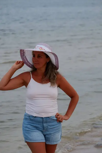 Tatil kumsalındaki kadın fotoğraf çekimi yapıyor farklı pozlar ve mekanlarda gülümsüyor sarı saçlı hasır şapka farklı çekim seçenekleri sunuyor güneşlenmeler deniz ve kumda yürüyor.