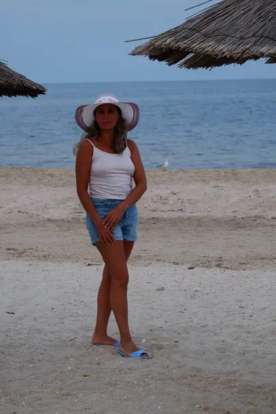 Tatil kumsalındaki kadın fotoğraf çekimi yapıyor farklı pozlar ve mekanlarda gülümsüyor sarı saçlı hasır şapka farklı çekim seçenekleri sunuyor güneşlenmeler deniz ve kumda yürüyor.