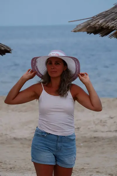 Tatil kumsalındaki kadın fotoğraf çekimi yapıyor farklı pozlar ve mekanlarda gülümsüyor sarı saçlı hasır şapka farklı çekim seçenekleri sunuyor güneşlenmeler deniz ve kumda yürüyor.