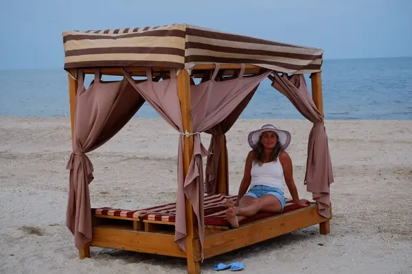 Tatil kumsalındaki kadın fotoğraf çekimi yapıyor farklı pozlar ve mekanlarda gülümsüyor sarı saçlı hasır şapka farklı çekim seçenekleri sunuyor güneşlenmeler deniz ve kumda yürüyor.