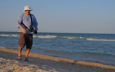 Orta yaşlı bir adam yazın deniz kıyısında yürüyor, kamera ile ve uzaktaki doğanın güzelliğinin fotoğraflarını çekiyor insanların deniz bulutları 1 'i görebiliyorsunuz.