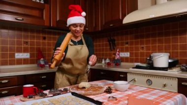 Noel Baba şapkalı ve bej renkli şef önlüklü güzel bir kadın mikrofon gibi merdaneye şarkı söylüyor, eğleniyor, evde geleneksel bir Noel pastası ve zencefilli kurabiye hazırlarken mutluluğu yaşıyor.