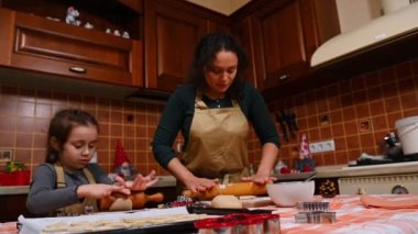 Güzel bir Latin Amerikalı kadın, sevgi dolu bir anne ve bej rengi önlüklü tatlı kızı, hamuru yuvarlıyor, zencefilli kek pişiriyor ve eski moda tasarımlarla mutfakta Noel pastası pişiriyor.