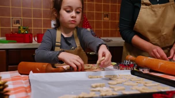 ベージュのシェフエプロンの素敵な娘である白人の小さな子供の女の子は 母親が自宅のキッチンでクリスマスペストリーを調理するのを助けます テーブルの上の羊皮紙に生地の型をカットします お祭り料理 — ストック動画