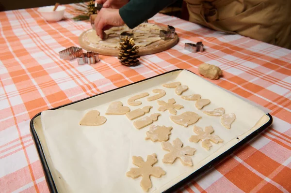 A baking sheet with gingerbread cookies from various shape on the parchment, on the background of a housewife confectioner cutting molds out of dough. Baking cookies for Christmas holidays. New Year