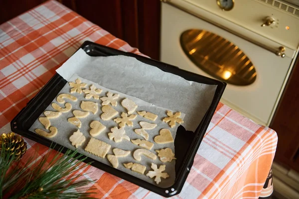 Top view of a baking sheet with cut-out gingerbread dough molds, on the kitchen table, against the background of a heating oven, in the wooden kitchen in vintage old design. Cozy Christmas atmosphere