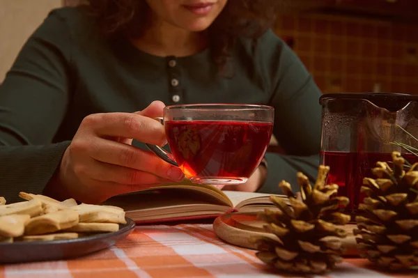 Selective focus on glass cup with hibiscus drink in the hand of a woman relaxing while reading book during tea time with gingerbread cookies. Golden pine cones as Christmas ornament on the foreground