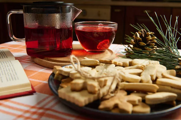 Hot winter drink: Hibiscus tea in a teapot and homemade gingerbread cookies on a plate. Christmas time. Cozy home atmosphere. Fir-tree branch and golden pine cones as decor. Holiday mood in the air