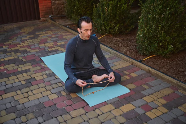 Top view of caucasian sportsman, yogi, sitting barefoot in lotus position on yoga mat, meditating with rosary. Sportsman doing breathing exercises outdoors using prayer beads. Body relaxing exercises