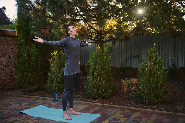 stock image Full-length of a Caucasian man yogi, an athlete in activewear outstretching arms palms up to the sky, standing barefoot on the yoga mat, deeply breathing, raising his body to the light at sunset