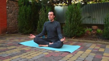 Güneşin batışı arka bahçeye vururken, yakışıklı aktif bir adam, yoga minderinde, yoga pozisyonunda meditasyon yaparken, avuç içlerini bir arada tutuyor. Gri spor kıyafetli barışçıl adam derin temiz hava soluyor..