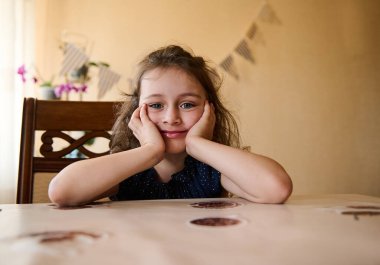 Sevimli beyaz bir çocuğun portresi, kıvırcık doğum günü kızı, tatlı tatlı gülümseyen, kameraya bakan, elleri yanaklarında masada oturan. Arka plandaki duvarda bayram bayrakları