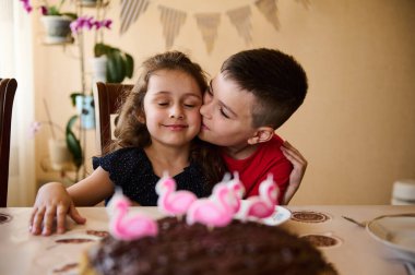Mutlu aile ilişkileri. Sevimli bir çocuk, sevgi dolu bir ağabey küçük kız kardeşine sarılıp öpüyor, bir doğum günü kızı, birlikte mutfak masasında oturuyor lezzetli bir ev yapımı çikolatalı şenlikli pastayla