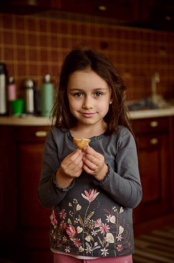 Güzel beyaz kız elinde zencefilli kurabiyeyle mutfakta dikilip kameraya bakarak tatlı tatlı gülümsüyor.
