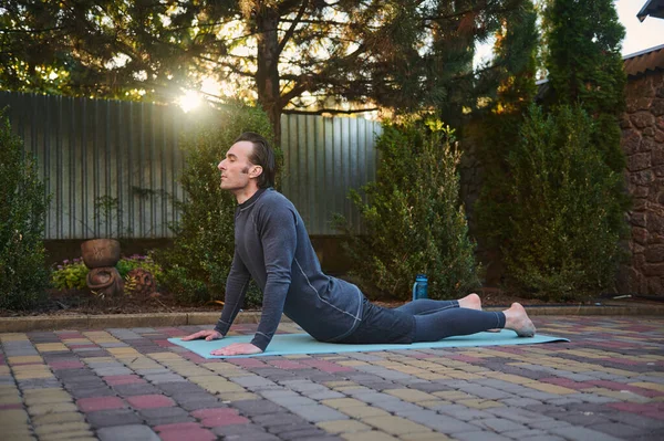 stock image Active sporty man athlete, a sportsman exercising on a fitness mat, doing stretching exercises outdoors at sunset. Sunbeams fall on the backyard while an athletic guy working out on the house backyard