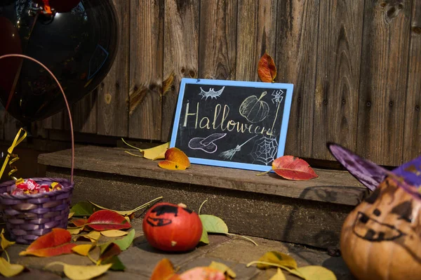 Halloween still life. Pumpkin in wizard hat, a purple basket with sweets ad candies next to a chalkboard and fallen autumn leaves on the wooden threshold. Halloween trick and treat