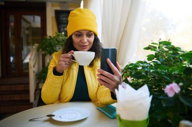 Parlak sarı şapka ve ceket giyen, Cafe 'de akıllı telefondan sohbet eden sevimli, mutlu Latin Amerikalı kadın. Güzel bir kadın güzel bir sonbahar gününde dışarıda kahve molası veriyor.