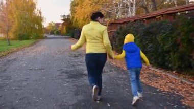 Mavi ve sarı ceketli mutlu bir anne ve kızın arka görüntüsü, güzel, sıcak bir sonbahar gününde kurumuş yaprakları olan bir caddenin ahşap çitleri boyunca birlikte koşarken el ele tutuşuyorlar.