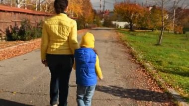 Sarı ceketli ve mavi kotlu esmer bir kadın, kızını ellerinden tutan sevgi dolu bir anne, güzel, güneşli bir sonbahar gününde sokakta birlikte yürüyor. Birlikte hafta sonu.