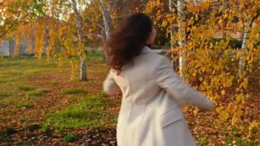 Gülümseyen güzel bir kadın, yeşil süveter ve bej, sıcak yün ceket giyiyor, hayattan zevk alıyor, düşen kuru portakal yapraklarını fırlatıyor, sonbahar parkında bir huş ağacının arka planında kıvrılıyor. Hint Yazı