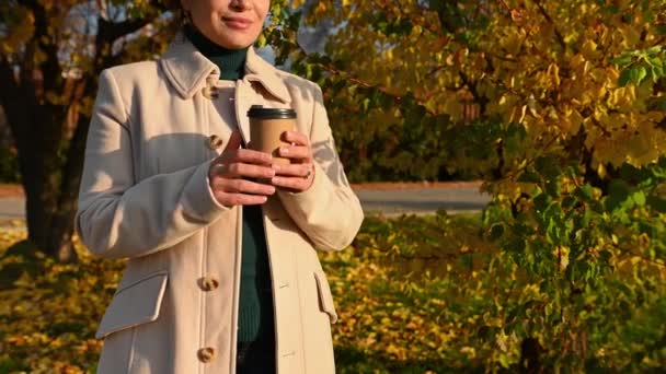 选择关注一位穿着米黄色外套的漂亮女人的手 用纸杯热咖啡加热她的手 同时享受生活和美丽而温暖的阳光普照的日子 漫步在秋天的公园 — 图库视频影像
