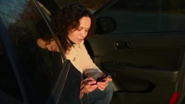 Güzel Latin Amerikalı kadın sürücü koltuğunda oturuyor ve akıllı telefon kullanıyor, haber yayını yapıyor, sosyal medyadaki içeriği kontrol ediyor, web sitelerini araklıyor. Sonbahar günbatımı güneş ışığı yüzüne vuruyor.