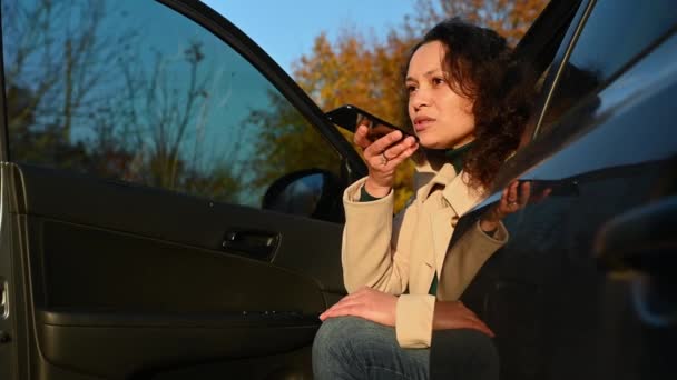 夕阳西下落在一位迷人的黑发多民族女性的脸上 她坐在停车场的司机座位上 在她的智能手机上记录着一条语音信息 — 图库视频影像