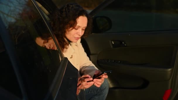美丽的拉丁美洲女人坐在司机座位上的汽车上 使用智能手机 滚动新闻资料 在社交媒体上查看内容 浏览网站 秋天的夕阳西下落在她脸上 — 图库视频影像