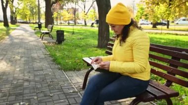 Sarı şapkalı, ceketli, tek kullanımlık kağıt bardaktan kahve içen, kitap okuyan, güneşli bir günde sonbahar parkında ahşap bir bankta oturan çok ırklı, rahat bir kadın. Kasım