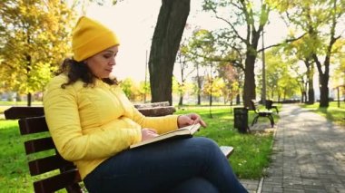 Parlak sarı ceketli ve şapkalı çok ırklı bir kadın güneşli bir günde, sonbahar parkında dinlenirken ahşap bir bankta oturmuş kitap okuyor. Dijital detoks, edebiyat. Boş zaman. Hint Yazı