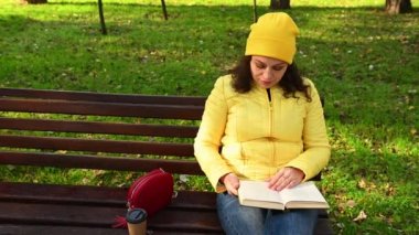 Mutlu, rahat bir kadın, sarı bir şapka ve ceket giymiş, haftasonunun keyfini çıkarıyor, kitap okurken sayfaları çeviriyor, üzerinde kağıt bardak içinde kahve olan tahta bir bankta oturuyor, sonbahar parkında