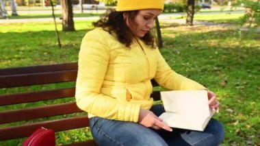 Sarı ceketli çekici bir kadın huş ağacının ve akçaağaç parkının ahşap bir bankında güneşli bir sonbahar gününde huzur dolu sakin bir atmosferde kitap okuyor ve Hint yazının son sıcak günlerinin tadını çıkarıyor.