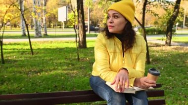 Sarı yün şapkalı, ceketli mutlu bir kadın, sonbahar parkında bir bankta oturuyor, düşünceli bir kenara bakıyor, kağıt bardaktan sıcak kahve içiyor, kitap okuyor.
