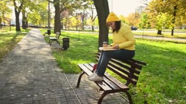Sarı şapkalı, ceketli, kağıt bardaktan kahve içen, kitap okuyan, güneşli bir günde, sonbahar parkında ahşap bir bankta oturan çok ırklı, hoş bir kadının düzenli bir fotoğrafı.