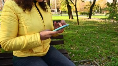Yakın plan kadın elleri akıllı telefondan haber yayını yapar, internette sörf yapar, sonbahar parkında sosyal medya içeriğini kontrol eder. Sarı ceketli bir kadının görüntüsü kesilmiş, cep telefonuyla iletişim kuruyor.