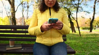 Sarı ceketli, ahşap bankta oturan, telefonuyla eğlenen, akıllı telefondan haber yayını yapan, sosyal medya içeriğini kontrol eden, çoklu etnik çeşitlilikte mutlu bir kadının ön görüntüsü.