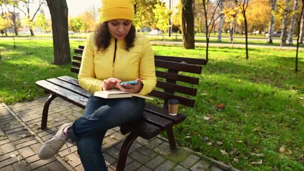 在一个阳光明媚的日子里 穿着鲜艳的黄色夹克 头戴一顶帽子的美丽而快乐的女人在秋天的公园里坐在长椅上 一边上网一边聊天 一边查看社交媒体的内容 — 图库视频影像