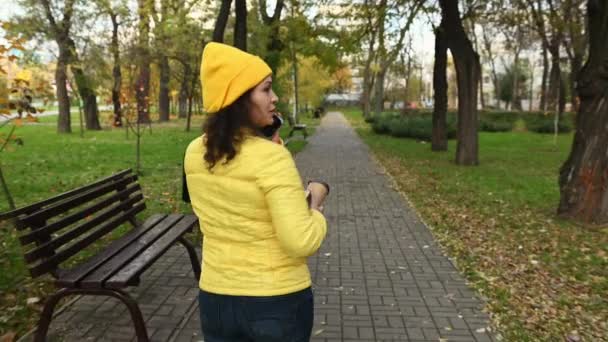 迷人的卷曲的多民族黑发女人 穿着鲜艳的黄色夹克 头戴羊毛帽 一边在秋天的公园边散步一边用手机聊天 手里拿着一本书和外卖食品 — 图库视频影像