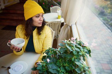 Parlak sarı şapkalı, ceketli, elinde beyaz bir fincan aromalı kahve tutan güzel bir kadının portresi, sonbahar parkında rahat bir kahve dükkanında dinlenirken pencereden dışarı bakıyor.