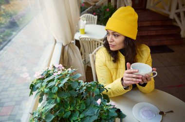 Milenyum Latin Amerikalı esmer kadın, sarı şapkalı ve ceketli, ellerini bir fincan sıcak aroma kahvesiyle ısıtıyor, sonbahar doğasına bakarak, rahat bir kahvede dinleniyor.